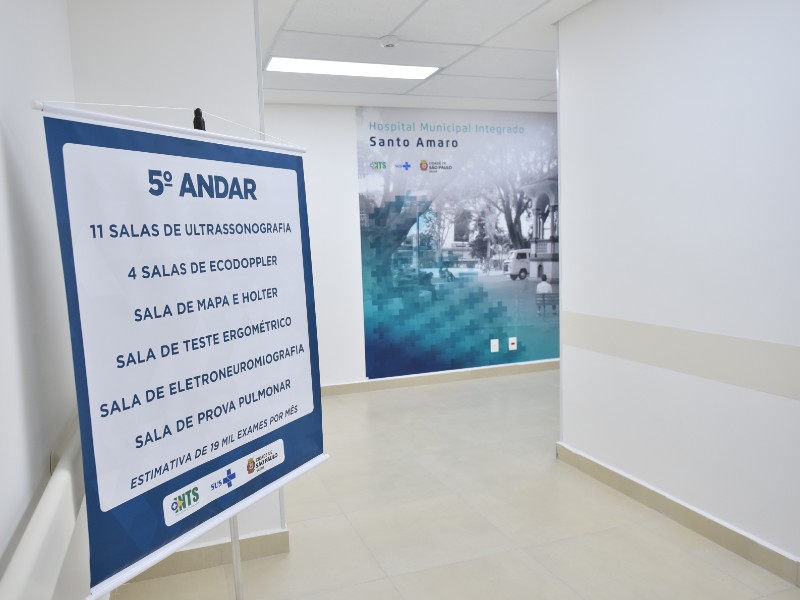 Corredor do hospital com paredes brancas e uma placa indicando o quinto andar da unidade. Ao fundo, um painel com foto em preto e branco e o nome do Hospital Municipal Integrado Santo Amaro impresso